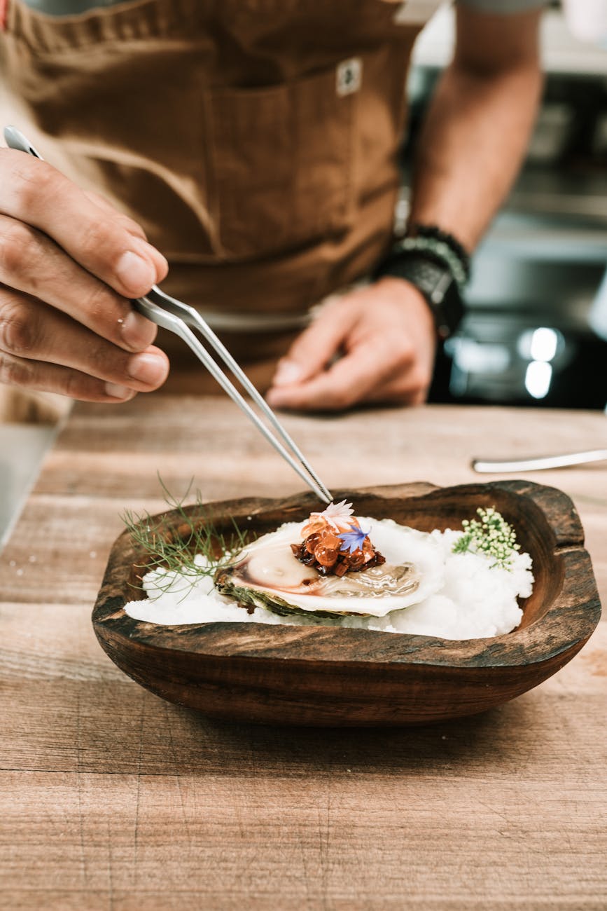 A chef delicately garnishing an oyster on a wooden plate with tweezers, showcasing a culinary presentation.